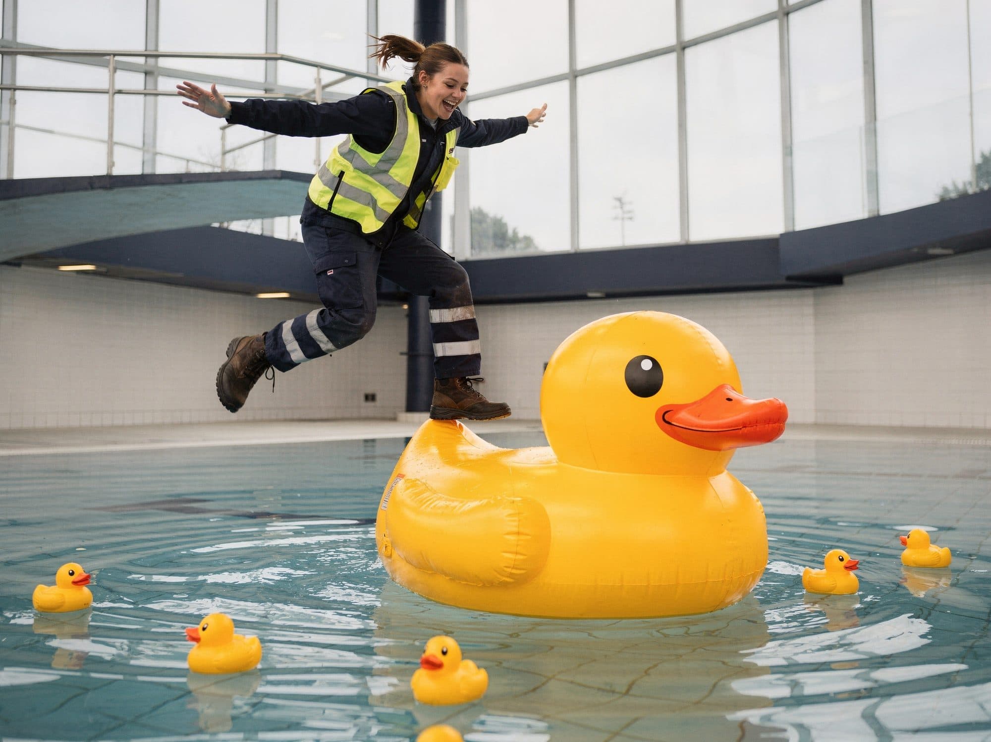 Laughing worker in workwear and a high-visibility vest jumps onto a giant inflatable rubber duck in an indoor pool, with small rubber ducks floating around